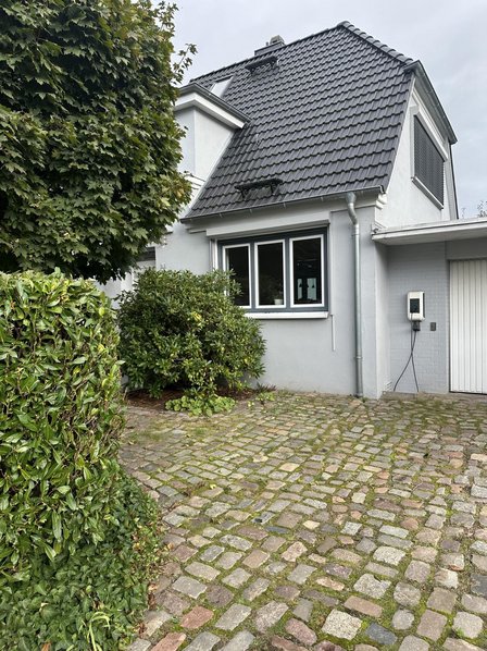 Hausfassade mit dunklem Ziegeldach, Einfahrt aus Kopfsteinpflaster und Sträuchern