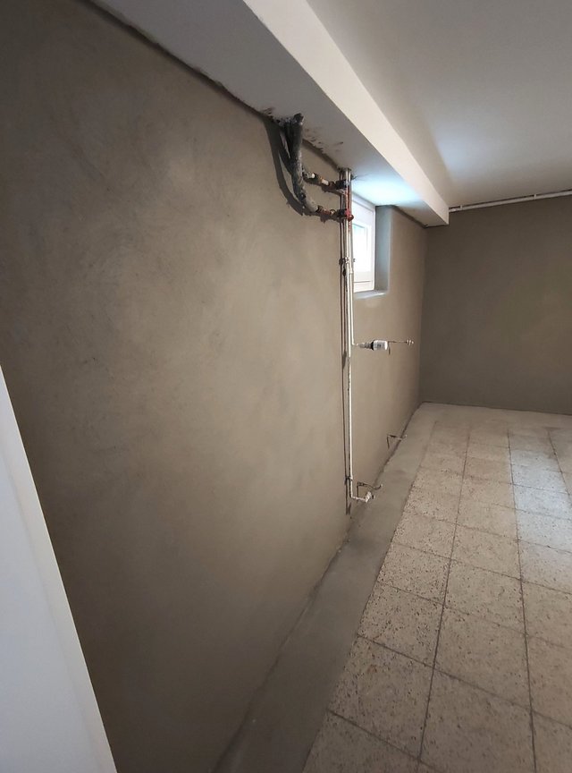 Kellerraum mit verputzter Wand, Heizungsrohren und kleinem Fenster neben Fliesenboden