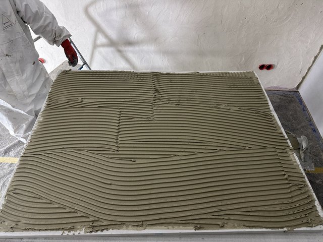 Arbeiter verteilt graue Masse gerillt auf heller Platte in einem Raum.