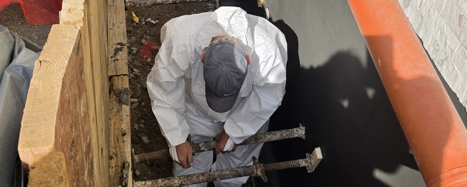 Arbeiter im Schutzanzug montiert Stahlstreben in schmalem Graben neben schwarzer Abdichtung.