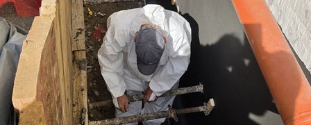 Arbeiter im Schutzanzug montiert Stahlstreben in schmalem Graben neben schwarzer Abdichtung.