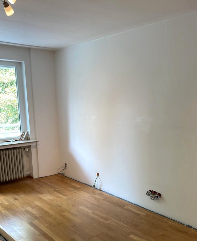 Leeres Zimmer mit Fenster, Heizkörper, Holzboden und freiliegenden Stromkabeln an der Wand