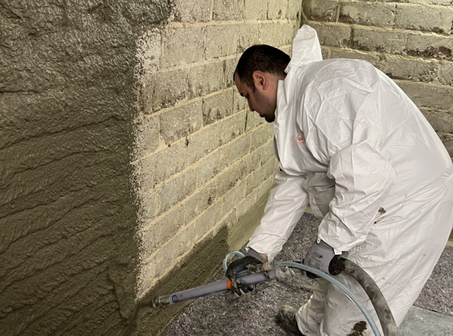 Arbeiter spritzt graue Masse auf Mauerwerk in einem Raum.