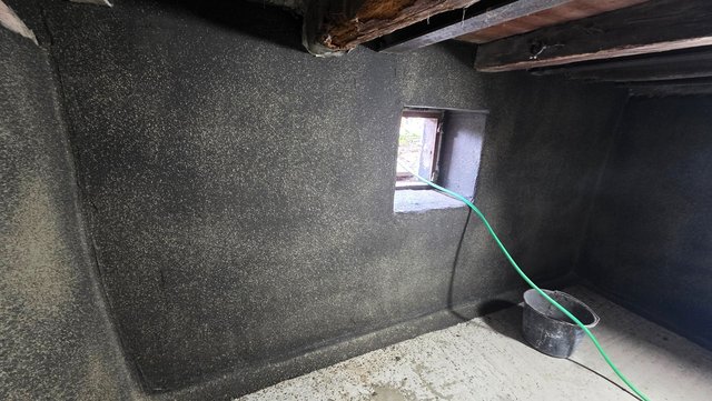 Schwarze Masse auf Kellerwand, Lichtschachtfenster und grüner Schlauch über Betonboden.
