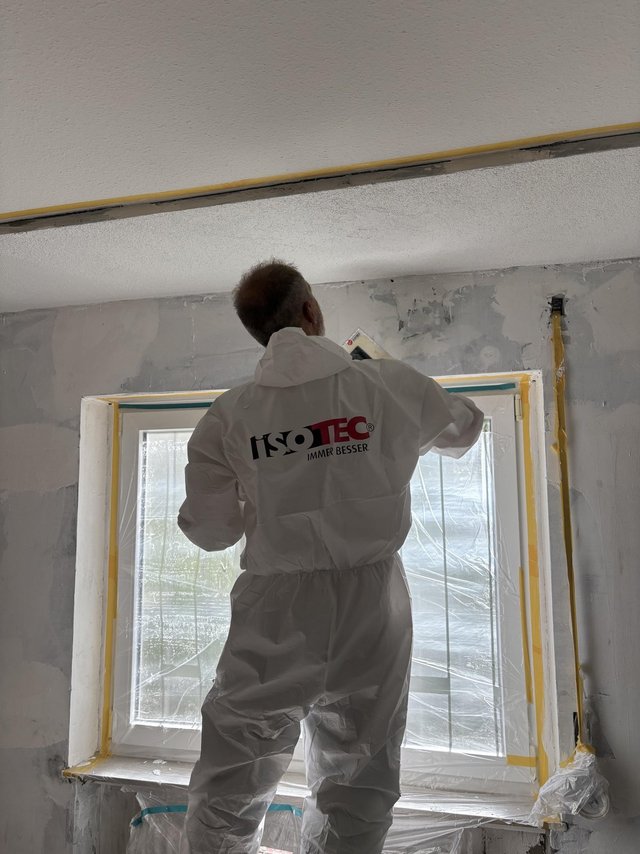 ISOTEC Mitarbeiter glättet graue Masse über Fensteröffnung, Folie und Klebeband schützen Rahmen.