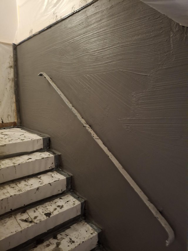 Treppenhauswand mit frisch aufgetragener grauer Masse und abgeklebtem Handlauf