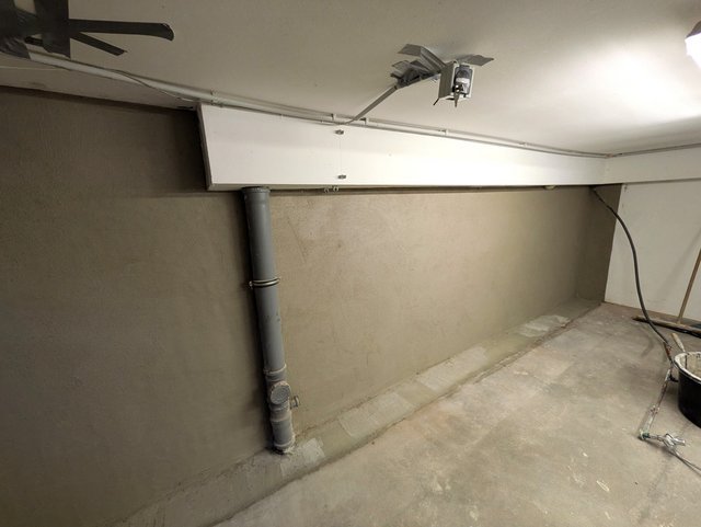 Abgedichtete Kellerwand mit grauer Masse, sichtbares Abflussrohr und Leitungen unter Decke