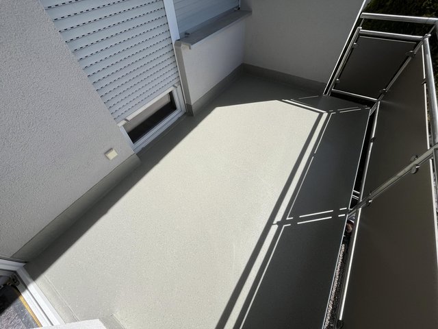 Sonniger Balkon mit hellgrauem Bodenbelag, Metallgeländer und Rollladenfenster