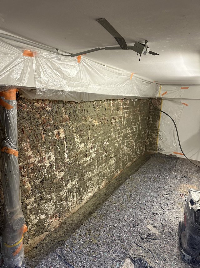 Freigelegte Kellerwand mit feuchten Ausblühungen, mit Folie abgeklebt und Baustellenboden.