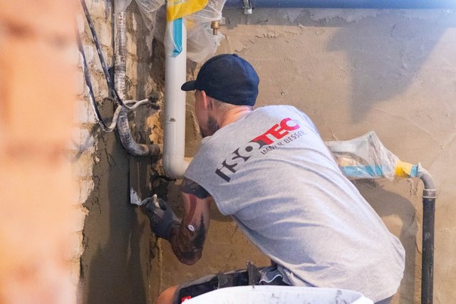 ISOTEC Mitarbeiter trägt graue Masse an Kellerwand neben Rohren auf