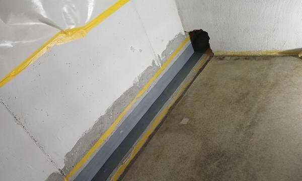Abgeklebter Wand-Boden-Übergang mit grauer Masse und schwarzem Streifen, Ecke mit Öffnung.