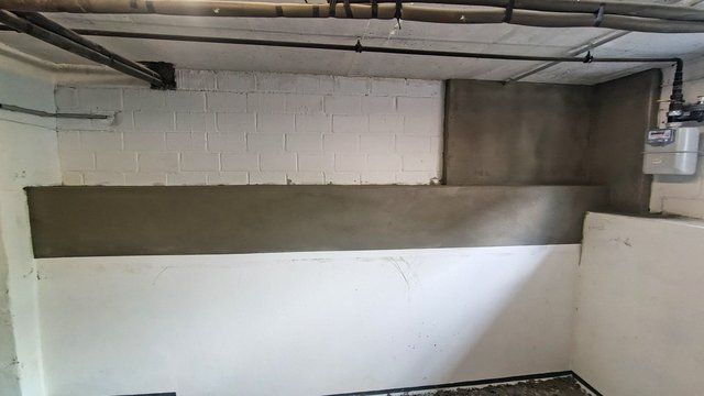 Kellerwand mit weißem Mauerwerk und aufgetragener grauer Masse, Deckenrohre darüber