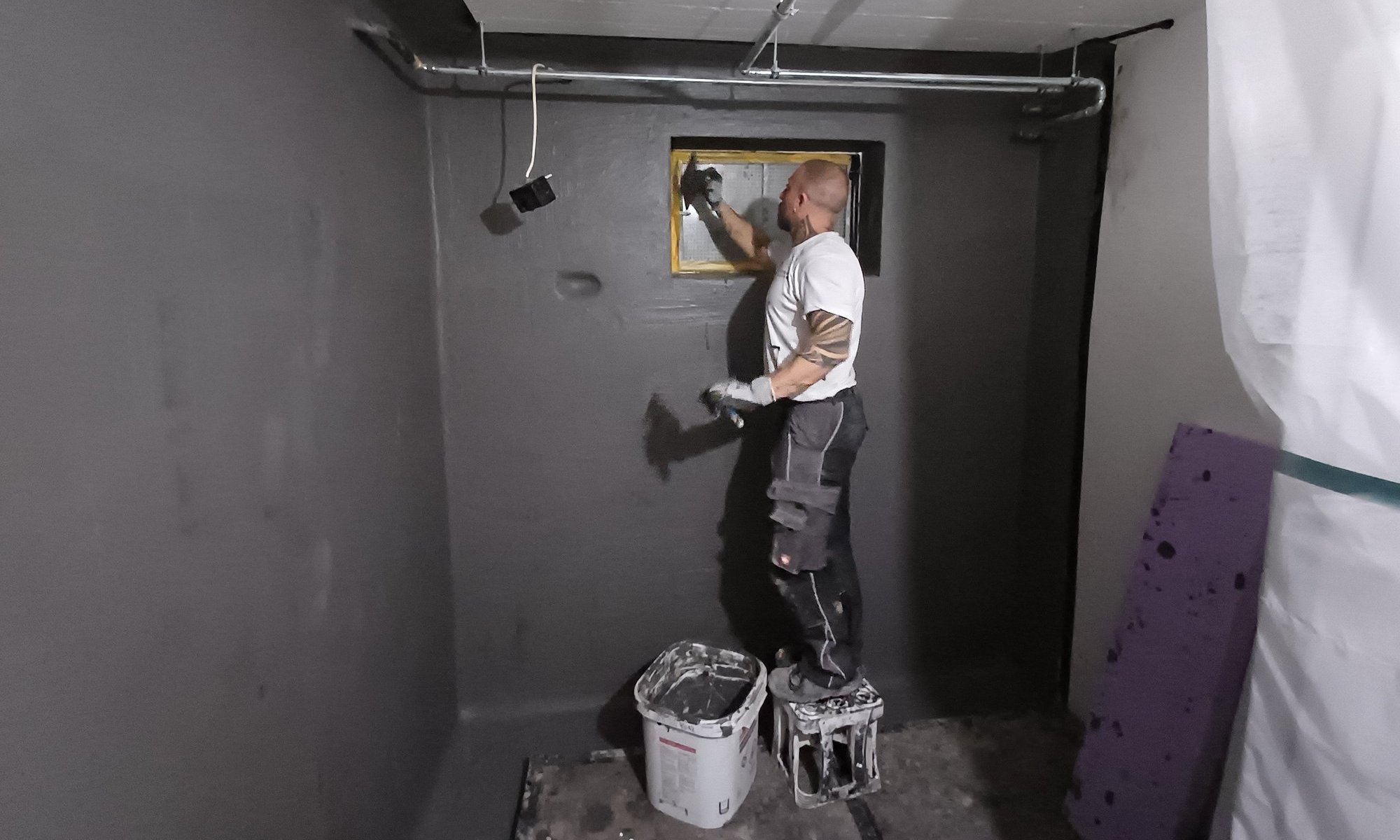 Arbeiter trägt schwarze Masse um Kellerfenster auf.
