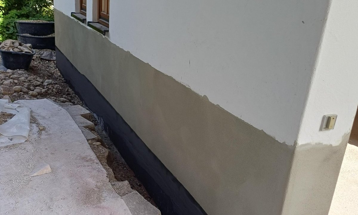Graue und schwarze Masse am Haussockel, freigelegter Graben entlang der Wand