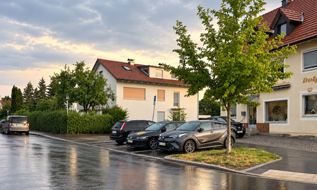 Nasse Straße mit parkenden Autos neben jungem Baum vor cremefarbenem Haus mit rotem Ziegeldach