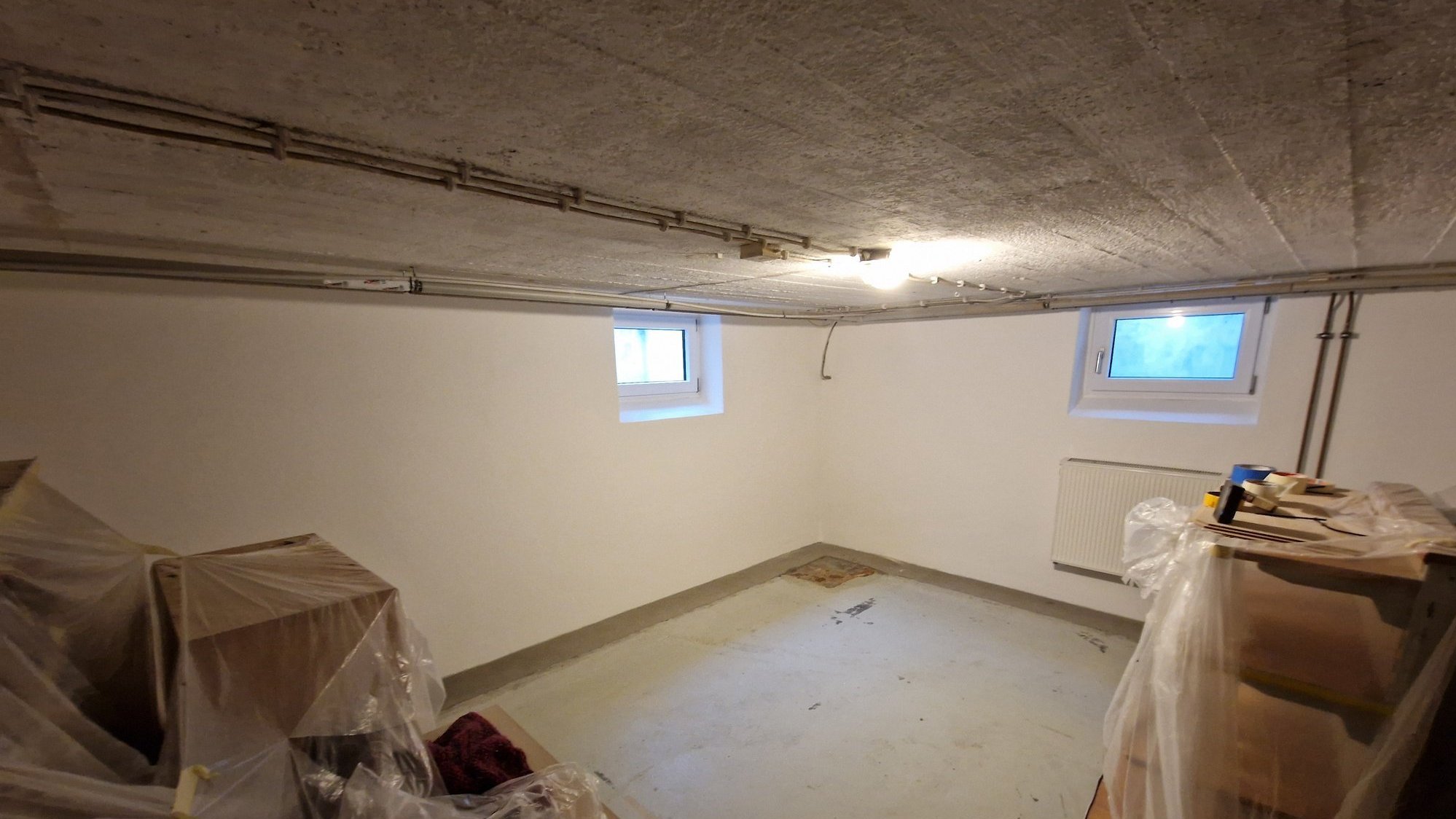 Leerer Kellerraum mit weißen Wänden, Beton­decke und abgedeckten Möbeln