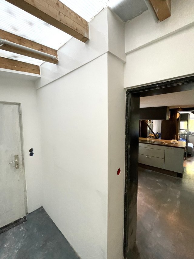 Weiß gestrichene Wand neben Holzbalken und Tür, Blick in Küche mit grauen Schränken