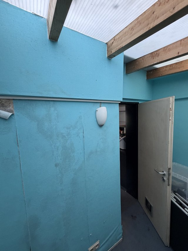Raum mit türkisfarbenen Wandpaneelen, fleckig, Holzbalken und Lichtplatten an der Decke, offene Tür