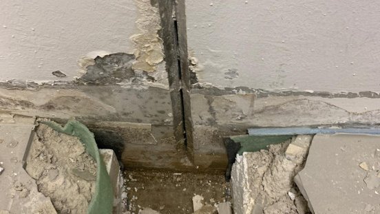 Hand zeigt freigelegten Wand‑Boden‑Übergang mit Riss und bröselndem grauem Material.