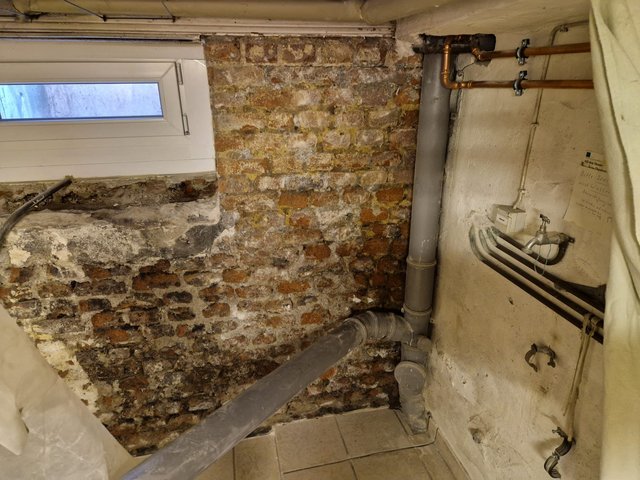 Freigelegte Kellerwand aus Mauerwerk mit Ausblühungen, Rohren und kleinem Fenster
