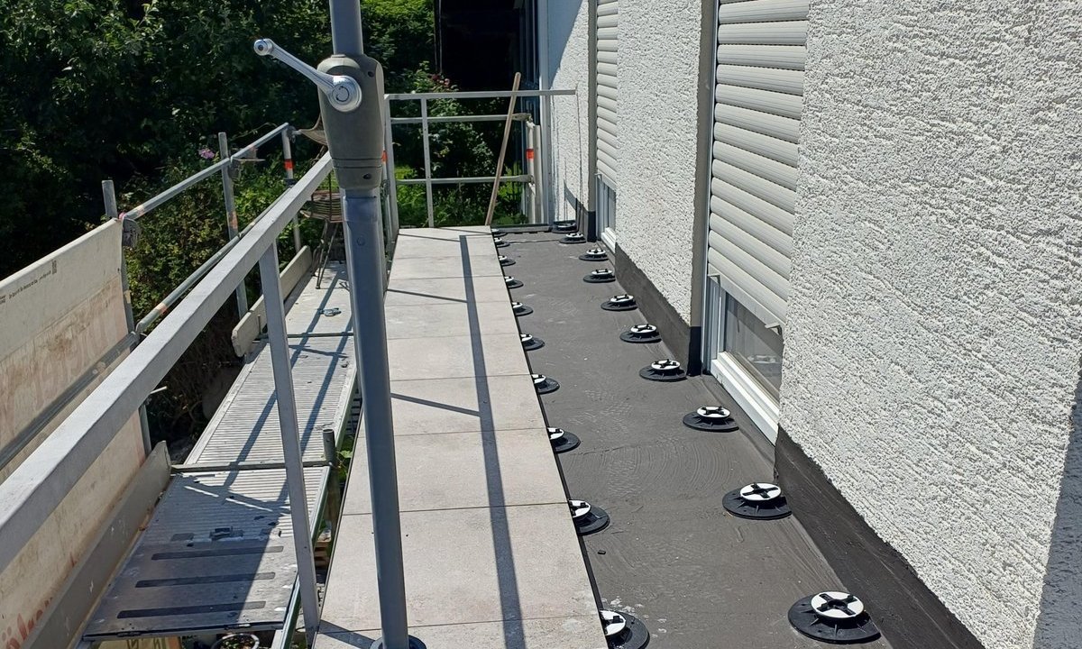 Balkon mit grauen Platten, schwarze Abdichtungsbahn und höhenverstellbare Stelzlager an der Hausfassade.