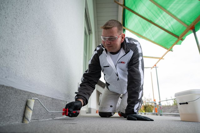 ISOTEC Mitarbeiter trägt helle Flüssigkeit mit Rolle auf Balkonboden entlang der Wand auf.