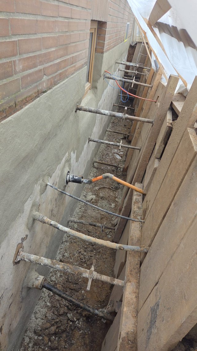 Freigelegte Hauswand im schmalen Erdgraben mit grauer Masse und Abstützungen