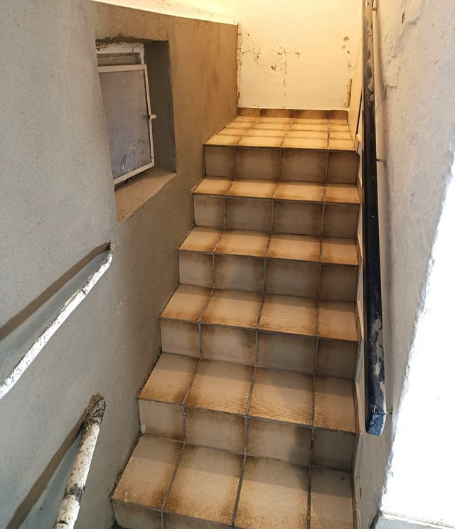 Geflieste Treppe in engem Treppenhaus mit verfärbten Wänden und kleinem Fenster
