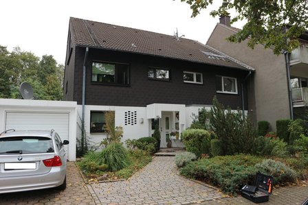 Einfamilienhaus mit dunkler Fassade, Garage, gepflasterter Einfahrt, parkendem Auto und Vorgarten.
