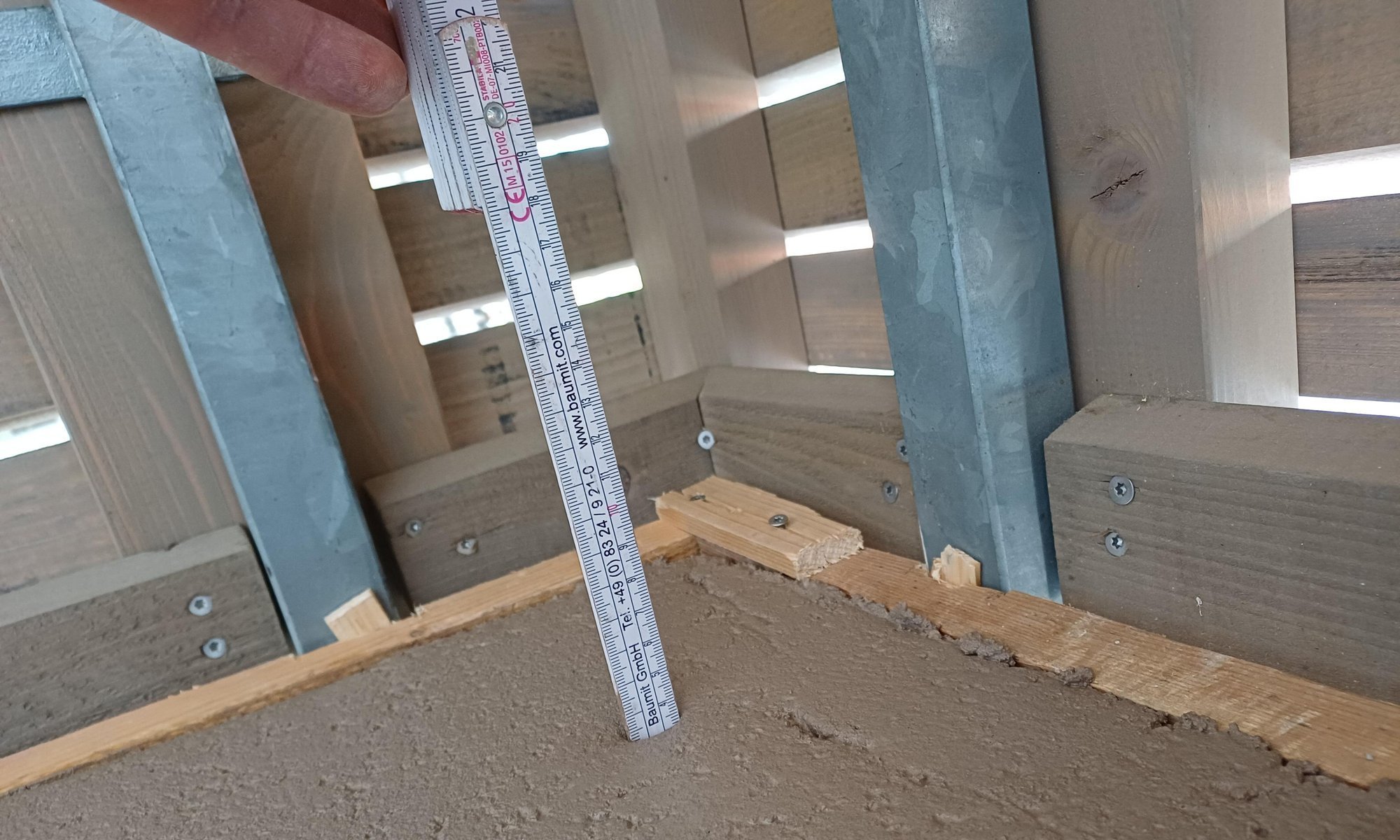 Hand misst mit Zollstock die Höhe der braunen Masse in einem Holzrahmen.