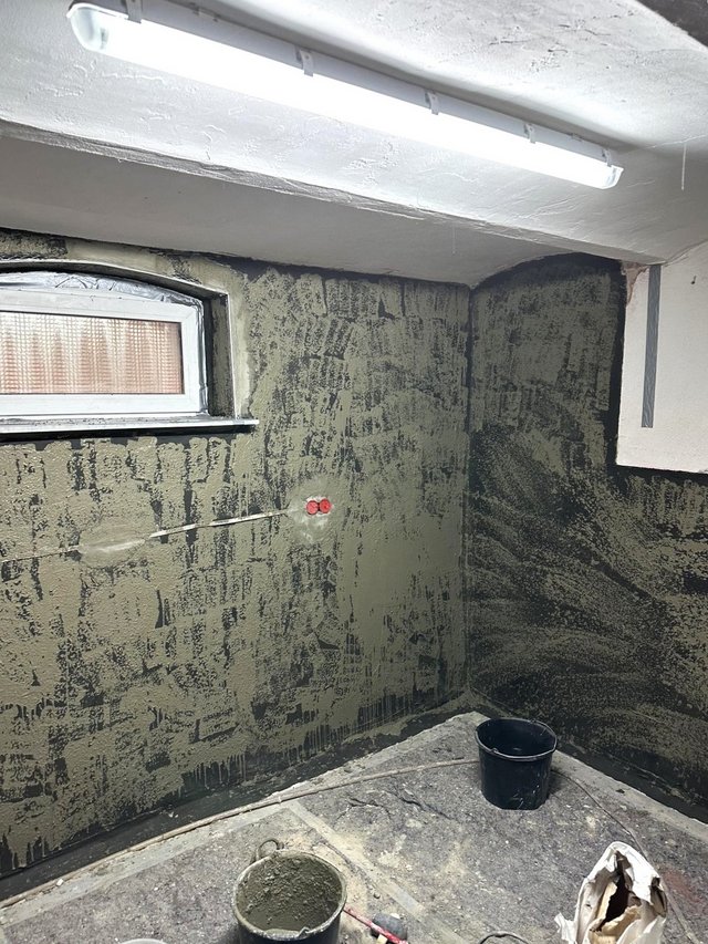 Graue Masse auf Kellerwand aufgetragen, Eimer und abgedeckter Boden im Raum