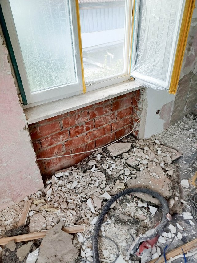 Rote Ziegelwand unter Fenster freigelegt, Bauschutt und Absaugschlauch liegen auf dem Boden.