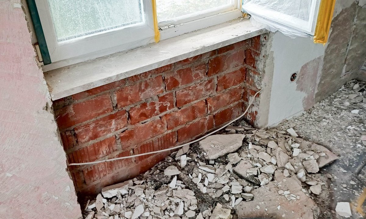 Rote Ziegelwand unter Fenster freigelegt, Bauschutt und Absaugschlauch liegen auf dem Boden.