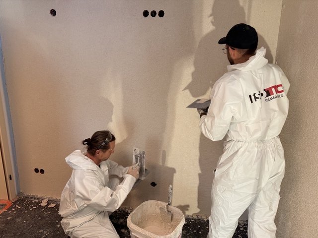 Zwei ISOTEC Mitarbeiter tragen graue Masse auf Wand auf.