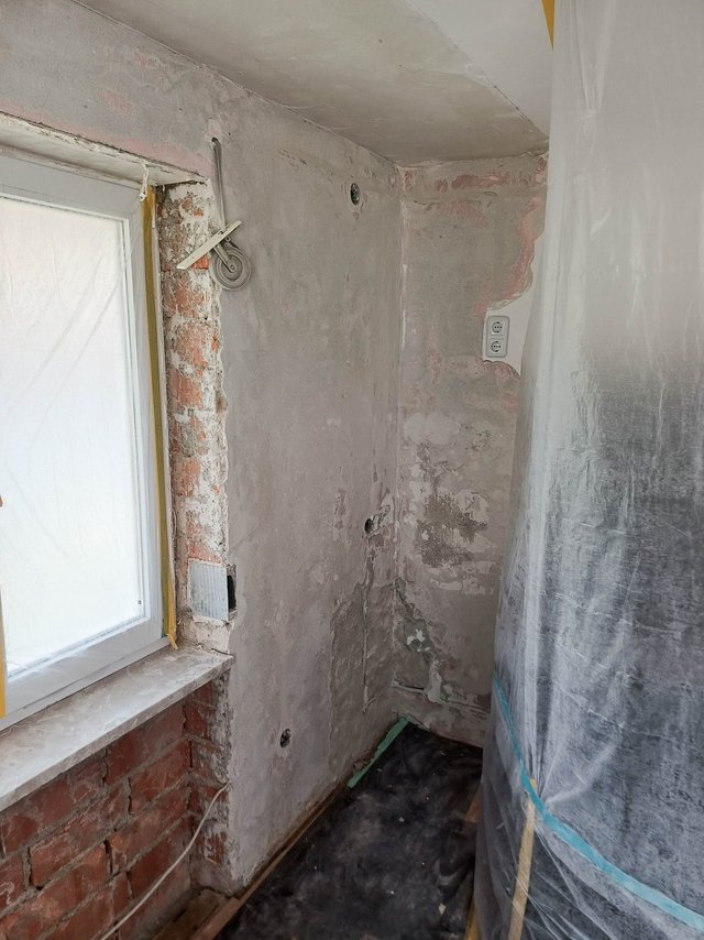 Rohbauraum mit grauer Masse an Wand, freigelegtes Ziegelmauerwerk neben Fenster, Schutzfolie an Seite