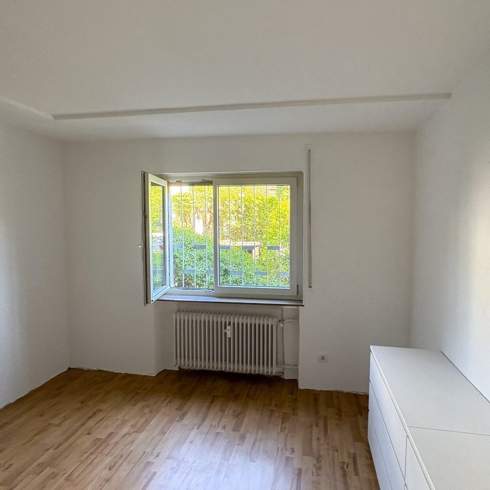 Leeres Zimmer mit Holzfußboden, geöffnetem Fenster mit Gitter und Heizkörper darunter