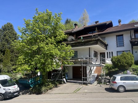 Wohnhaus mit Geruest am Balkon, gruener Baum im Vordergrund, zwei Autos am Strassenrand.