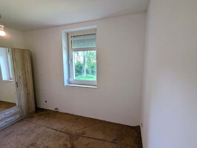 Leeres Zimmer mit weißer Wand, Fenster mit Rollladen und Holzschrank neben Spiegel