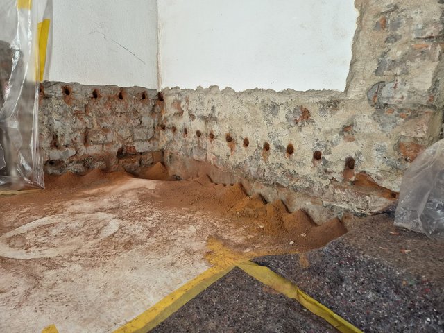 Bohrlöcher in freigelegtem Mauerwerk, Sandhäufchen am Boden, Baustellenabdeckung