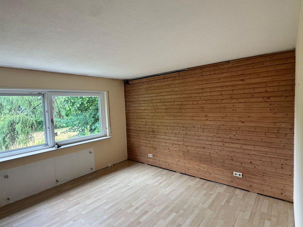Leeres Zimmer mit Fensterfront, Holzvertäfelung und hellem Laminatboden.