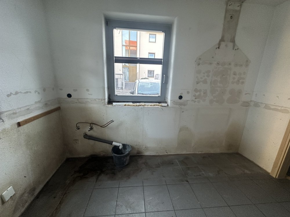 Leerer Raum mit Fenster, freigelegten Anschlüssen und Flecken an der Wand, Eimer am Boden.