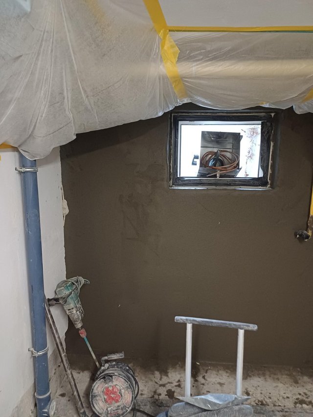 Abgeklebte Folienabdeckung an Decke, frische graue Masse um ein Kellerfenster.