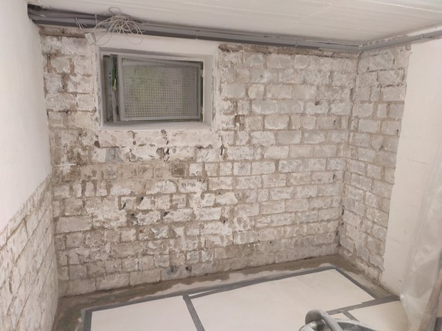 Freigelegte Kellerziegelwand mit kleinem Fenster und sichtbaren Kabeln an der Decke