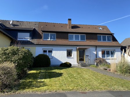 Einfamilienhaus mit braunem Ziegeldach, weißen Wänden und Vorgarten bei blauem Himmel
