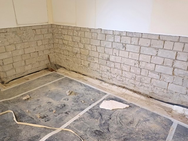 Schaufel lehnt an Wand mit grauer Masse, Boden ist mit schwarzer Folie abgedeckt.