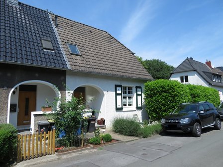 Weißes Wohnhaus mit Ziegeldach, grünen Fensterläden, Hecke und geparktem Auto.
