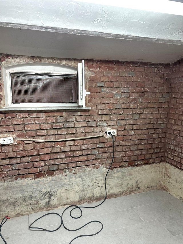 Freigelegte Ziegelwand mit Kellerfenster, Steckdosen und grauen Rückständen am Sockelbereich.