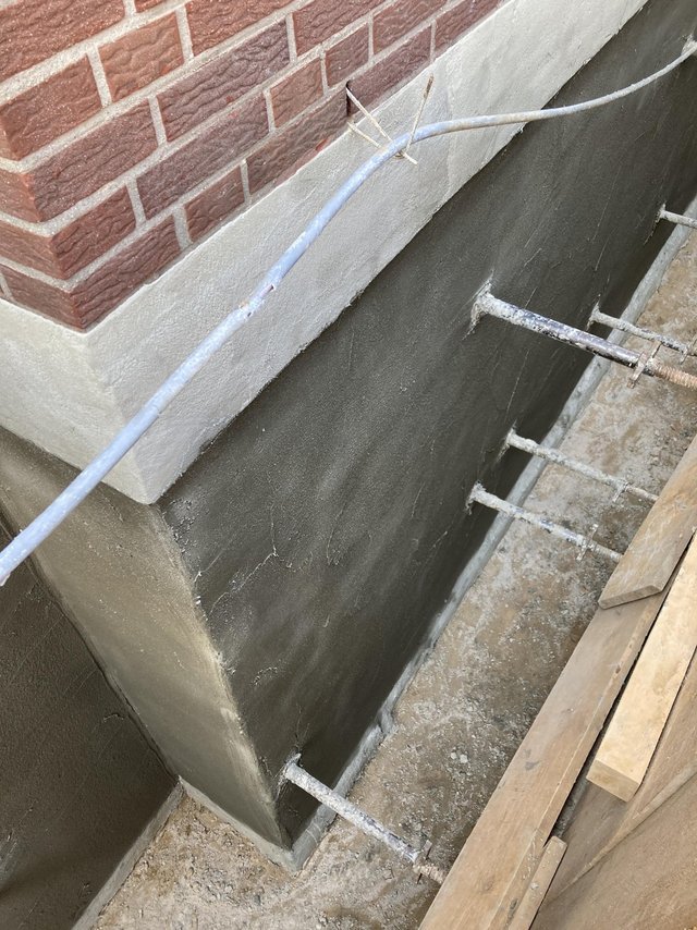 Schwarze Beschichtung auf Betonwand im Graben, Verankerungsstäbe ragen heraus.