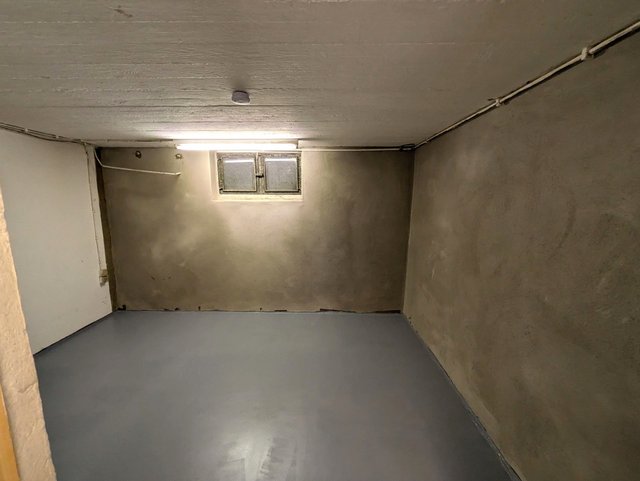 Leerer Kellerraum mit grauem Boden, verputzten Wänden und kleinem Fenster.