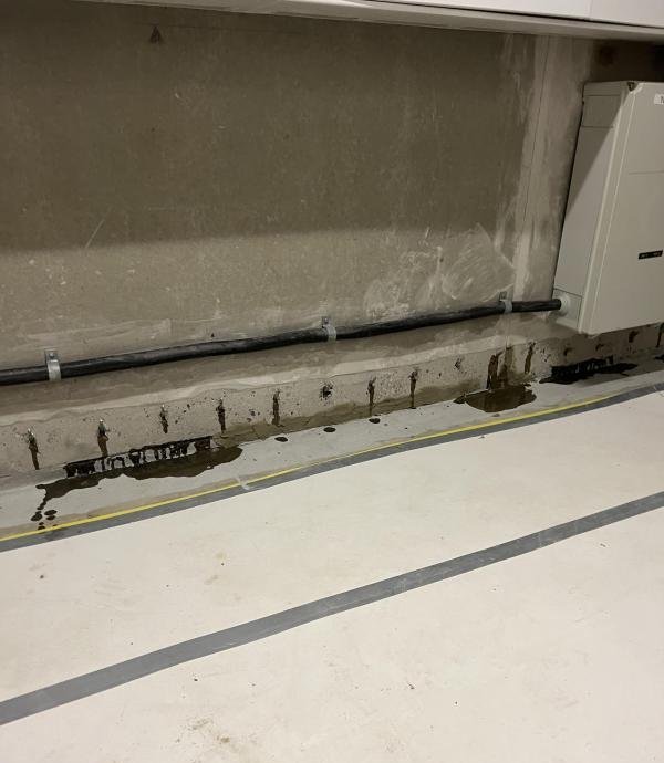 Dunkle Flüssigkeitsspuren am Boden entlang einer Betonwand mit Rohrleitung und Gerät